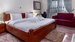 A neatly made bed with white linens and a towel shaped like swans, a wooden nightstand with a lamp, a red sofa, and a desk with a chair. - Hotel Green (Accra)