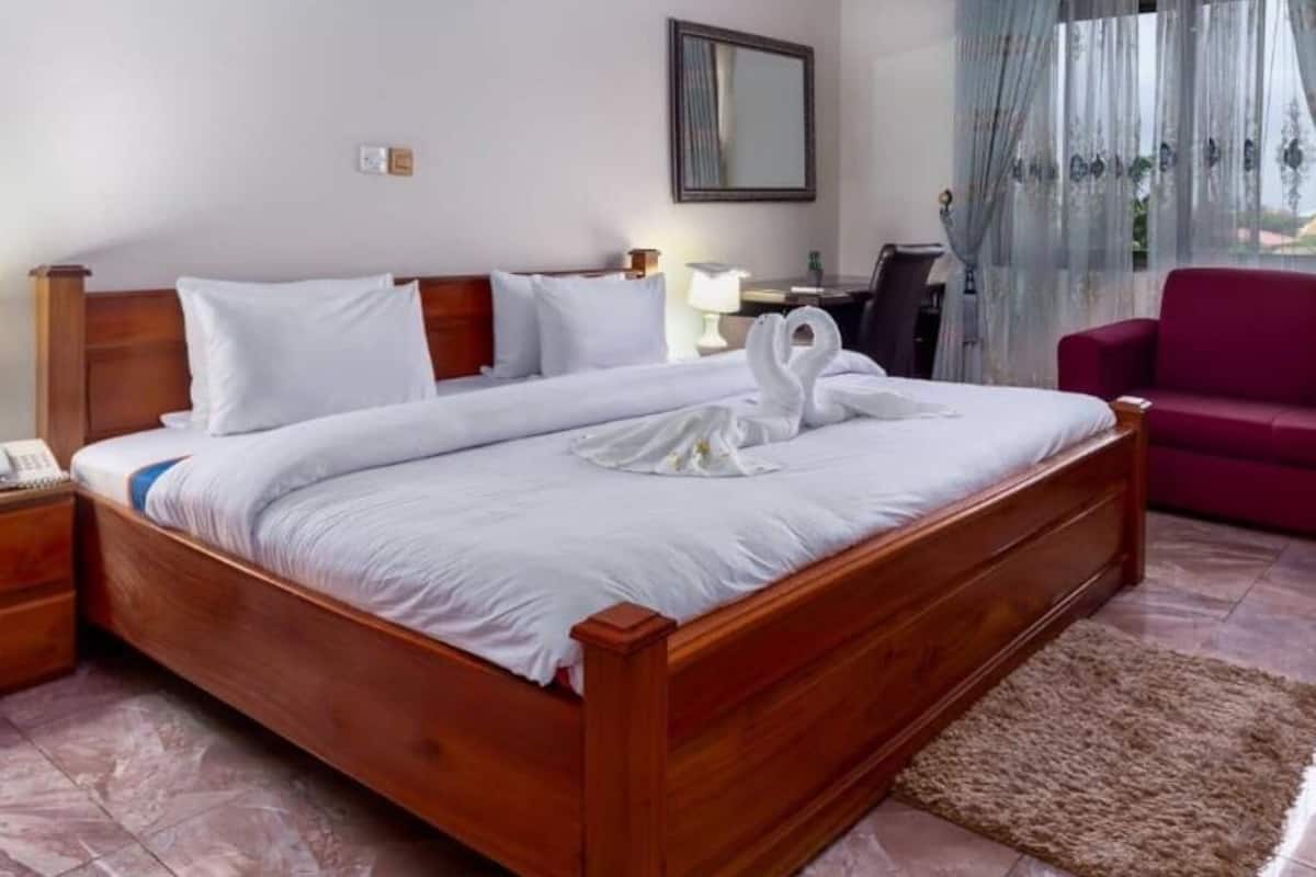 A neatly made bed with white linens and a towel shaped like swans, a wooden nightstand with a lamp, a red sofa, and a desk with a chair.