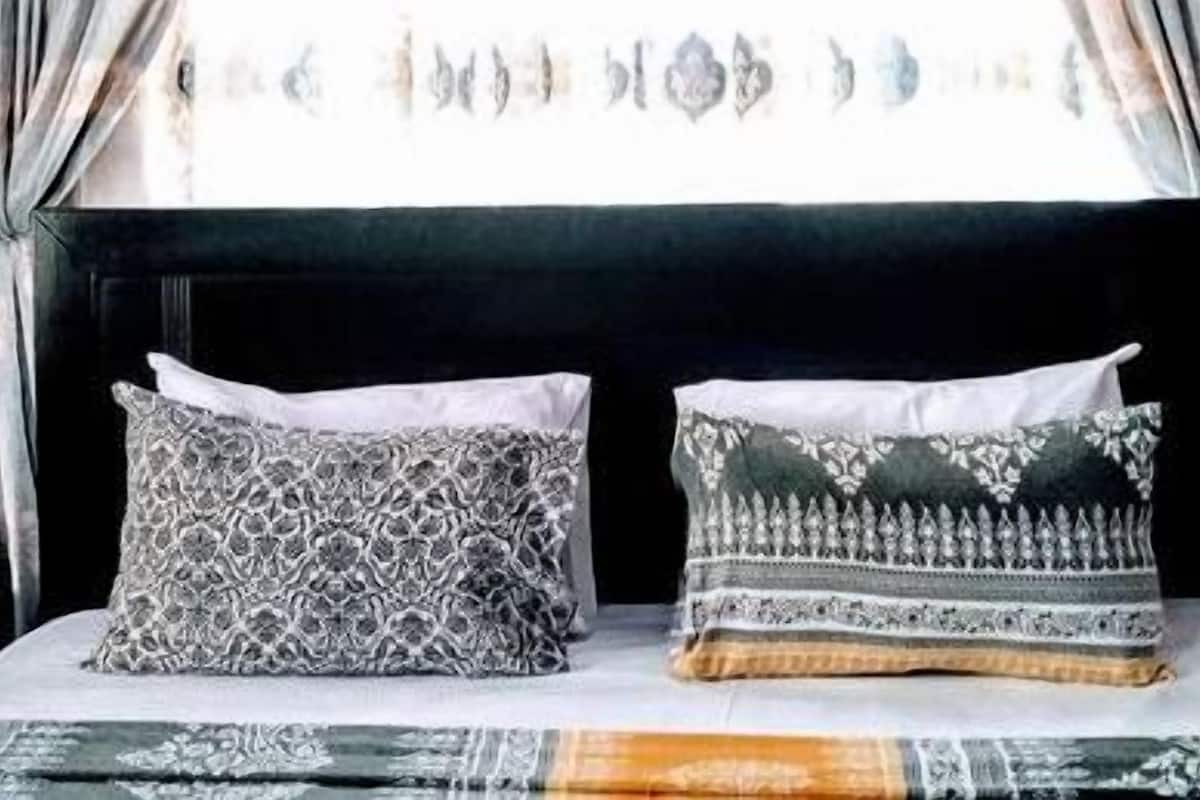 A bed with two patterned pillows, a bedside table with a lamp, and a window with curtains.