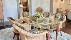 A dining area with a glass-top table surrounded by wicker chairs with leopard print cushions. There is a small side table with a plant and decorative items. The room has a carpeted floor and a doorway leading to another room. - Southern Staycation! (Greenville)