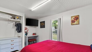 A bedroom with a red bedspread, a white dresser, a TV mounted on the wall, and a window with curtains.