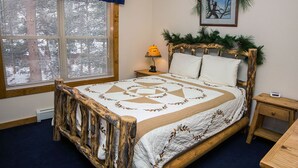 A bedroom with a wooden bed frame, a quilt, a nightstand with a lamp, and a window with blinds.