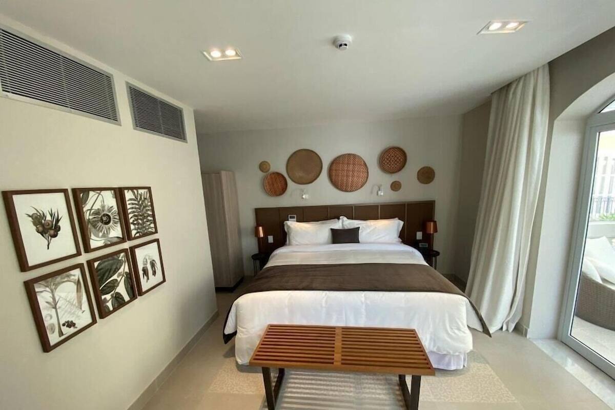 A modern hotel room with a large bed, a wooden bench, wall-mounted artwork, and a large window with curtains.