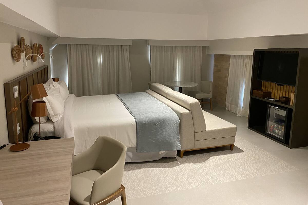 A modern hotel room with a large bed, a desk, a chair, a TV, and a minibar.
