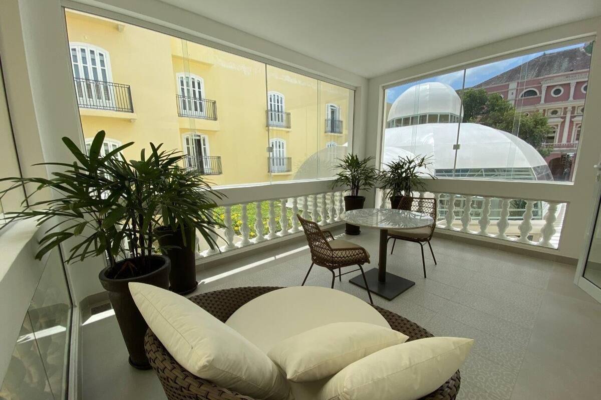 A balcony with wicker furniture, potted plants, and a view of yellow buildings.