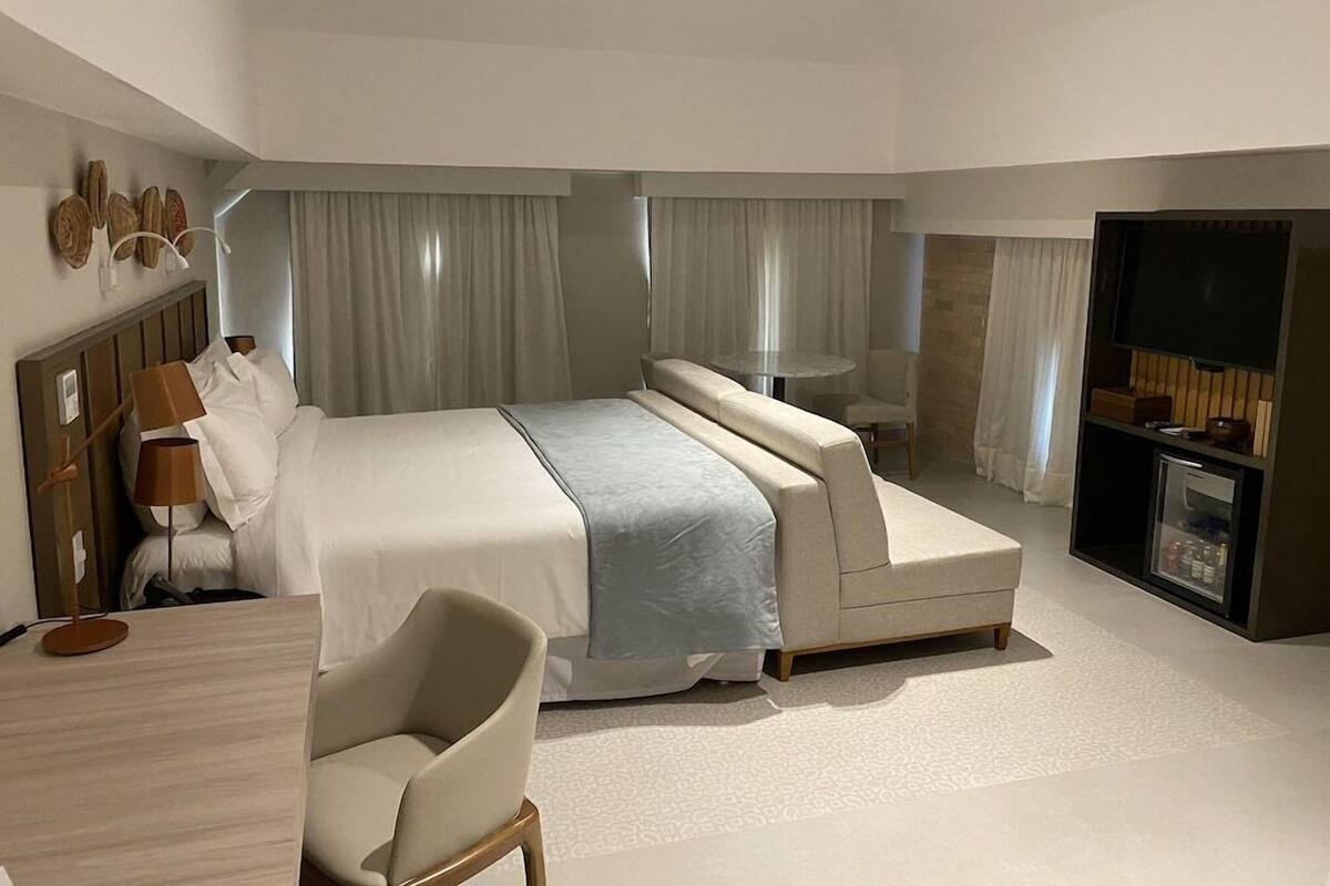 A modern hotel room with a large bed, a desk, a chair, a television, and a minibar.