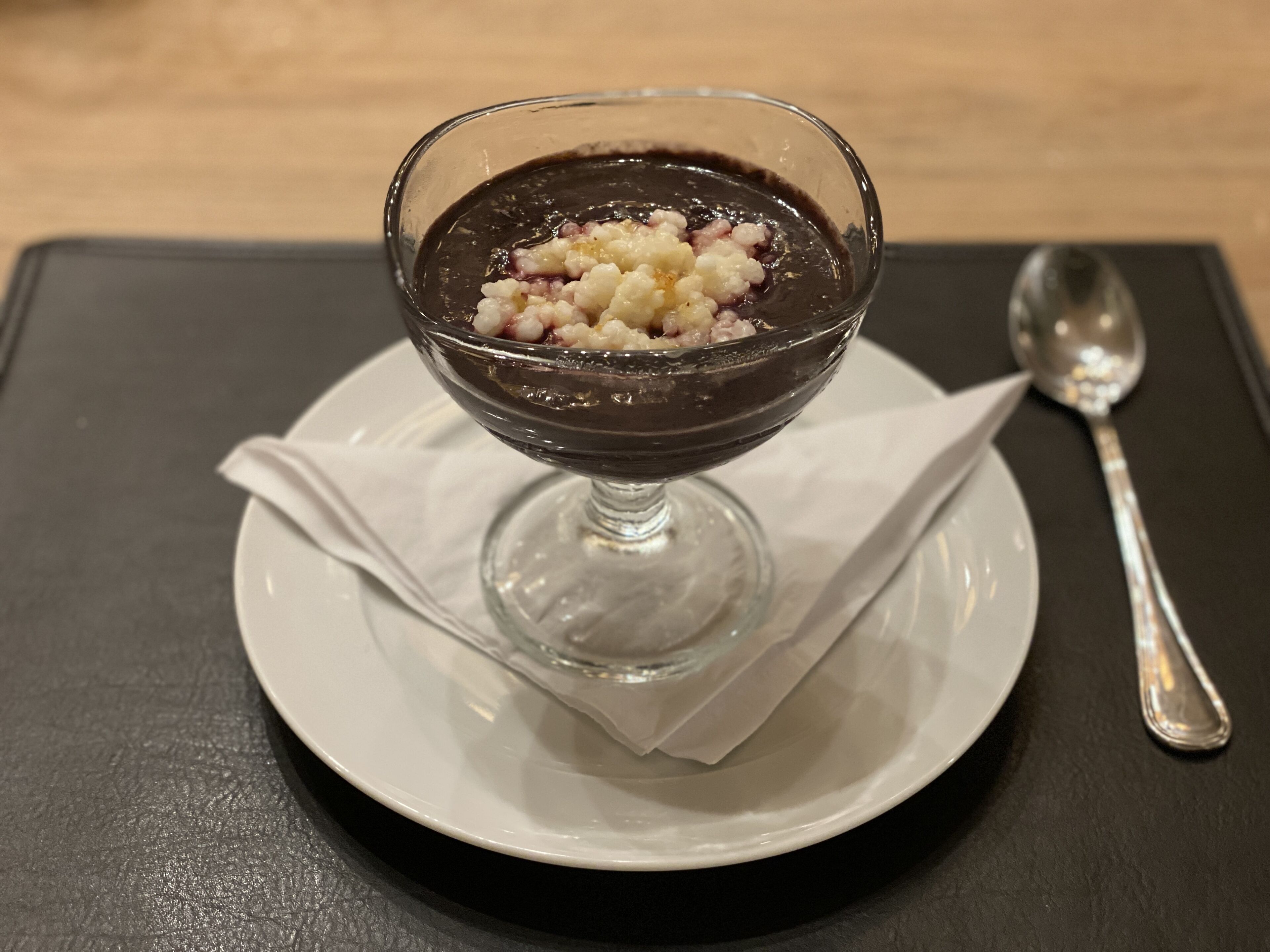 Un dolce in una ciotola di vetro su un piatto bianco, con un cucchiaio accanto.