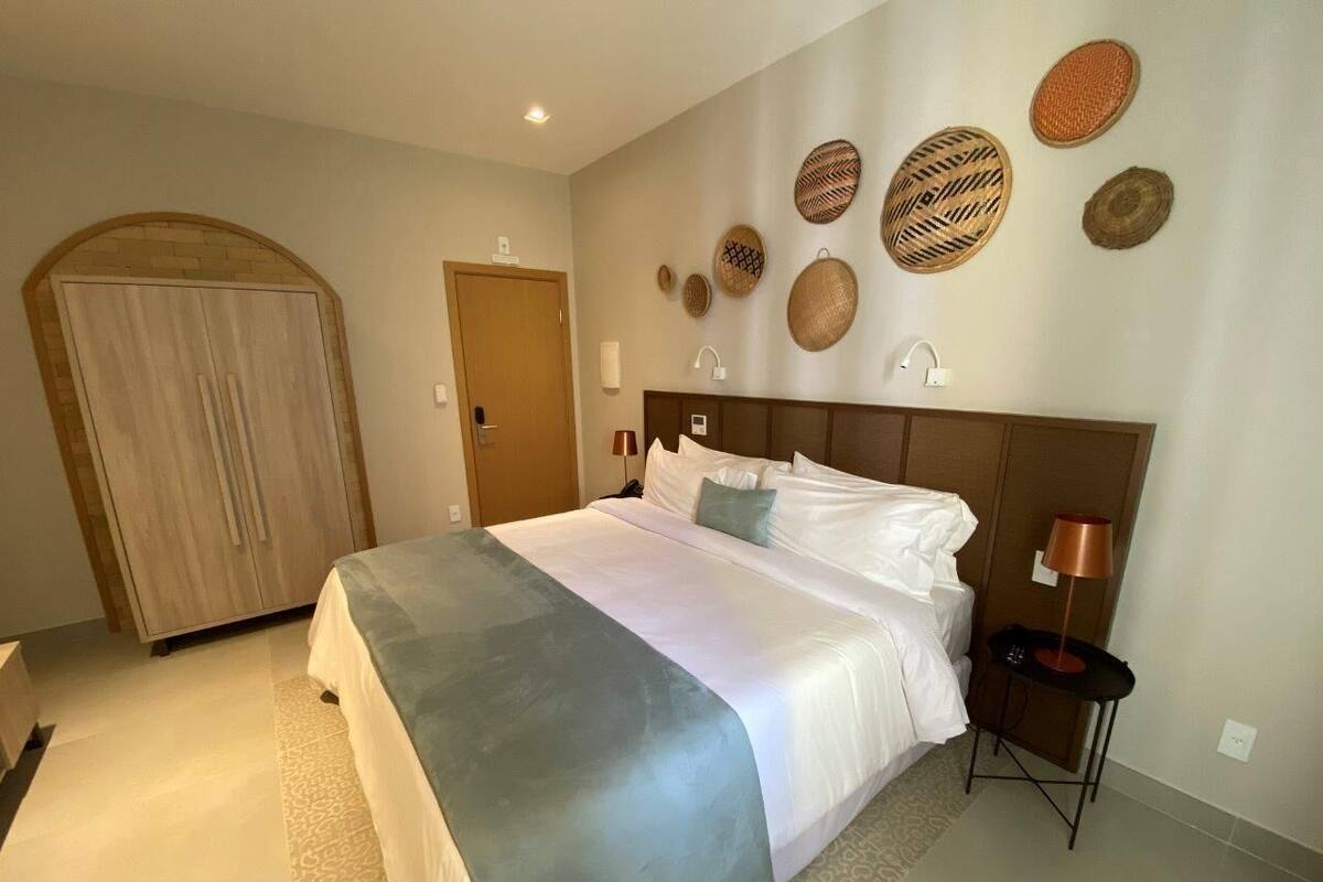 A hotel room with a large bed, a wardrobe, a bedside table with a lamp, and decorative woven baskets on the wall.