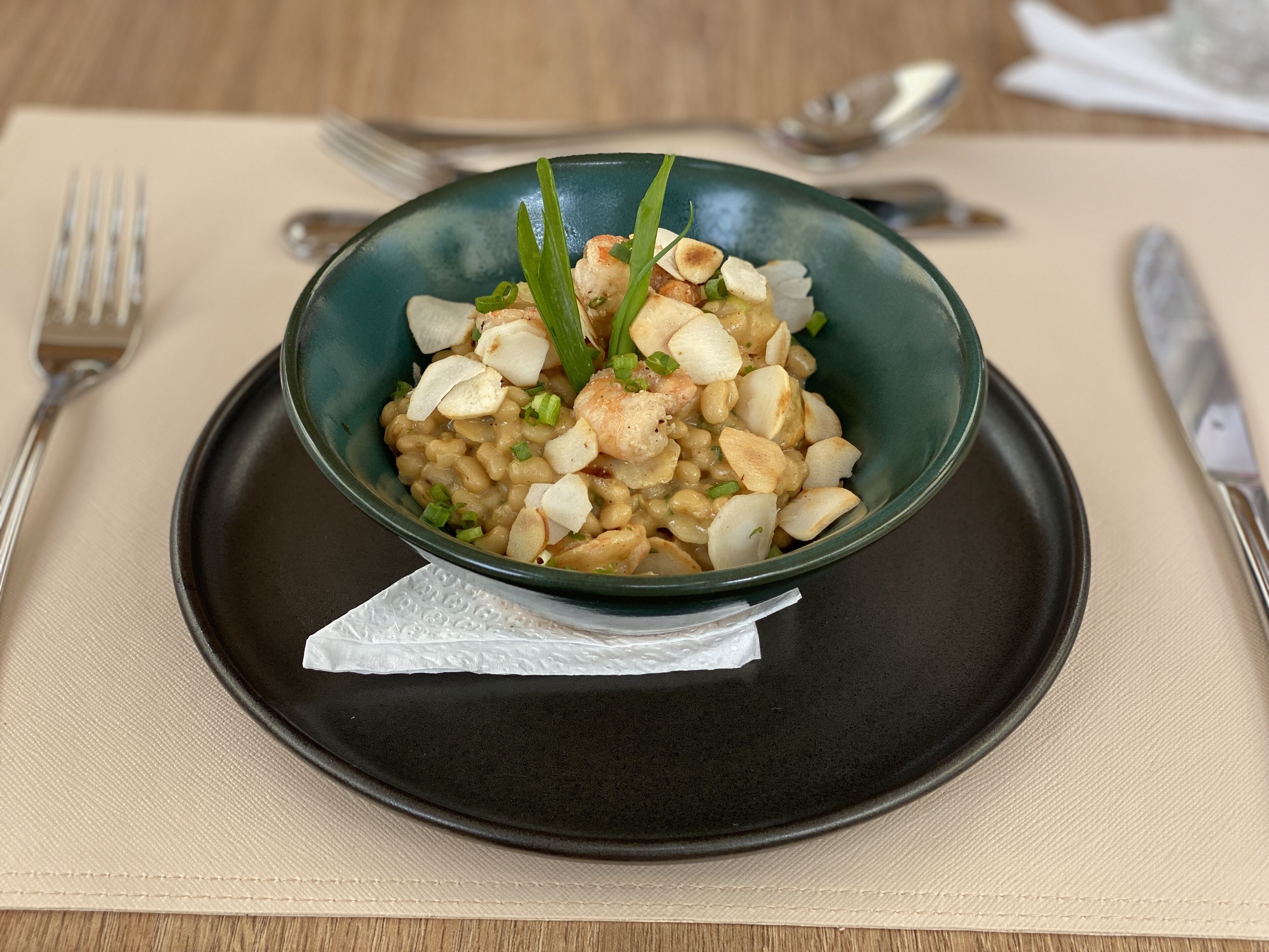 A bowl of food on a black plate with a napkin underneath, placed on a beige tablecloth.