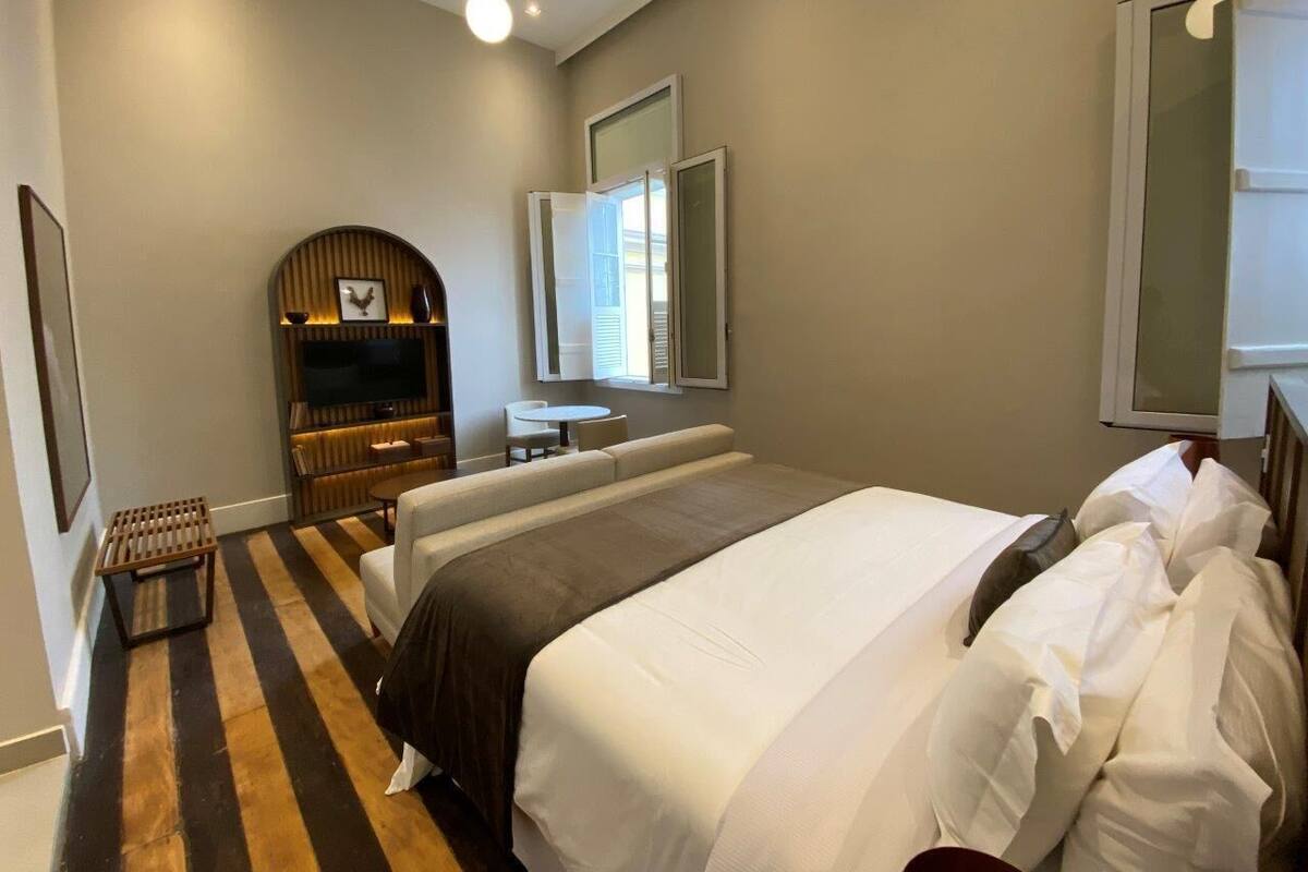 A bedroom with a large bed, a small table, a sofa, a TV, and a window with blinds.