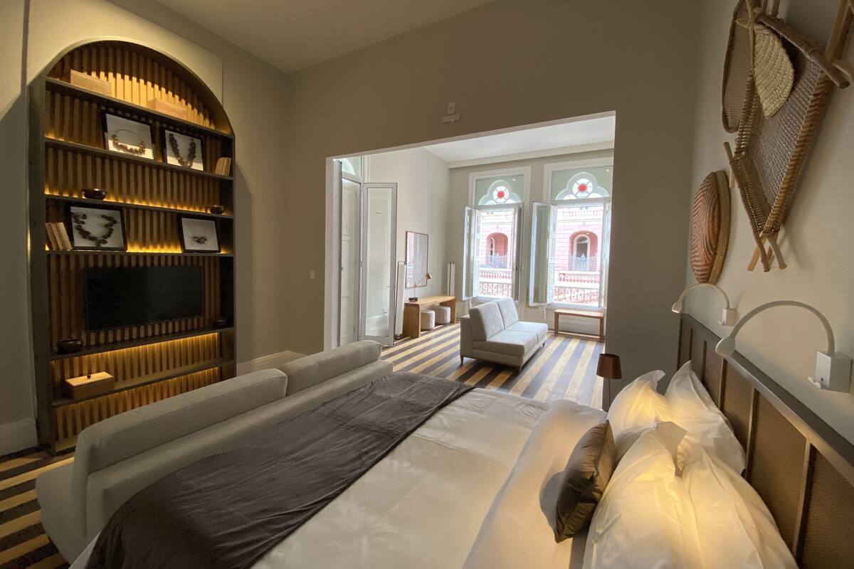 A modern hotel room with a large bed, a built-in bookshelf, a TV, and a view of a courtyard through a large window.