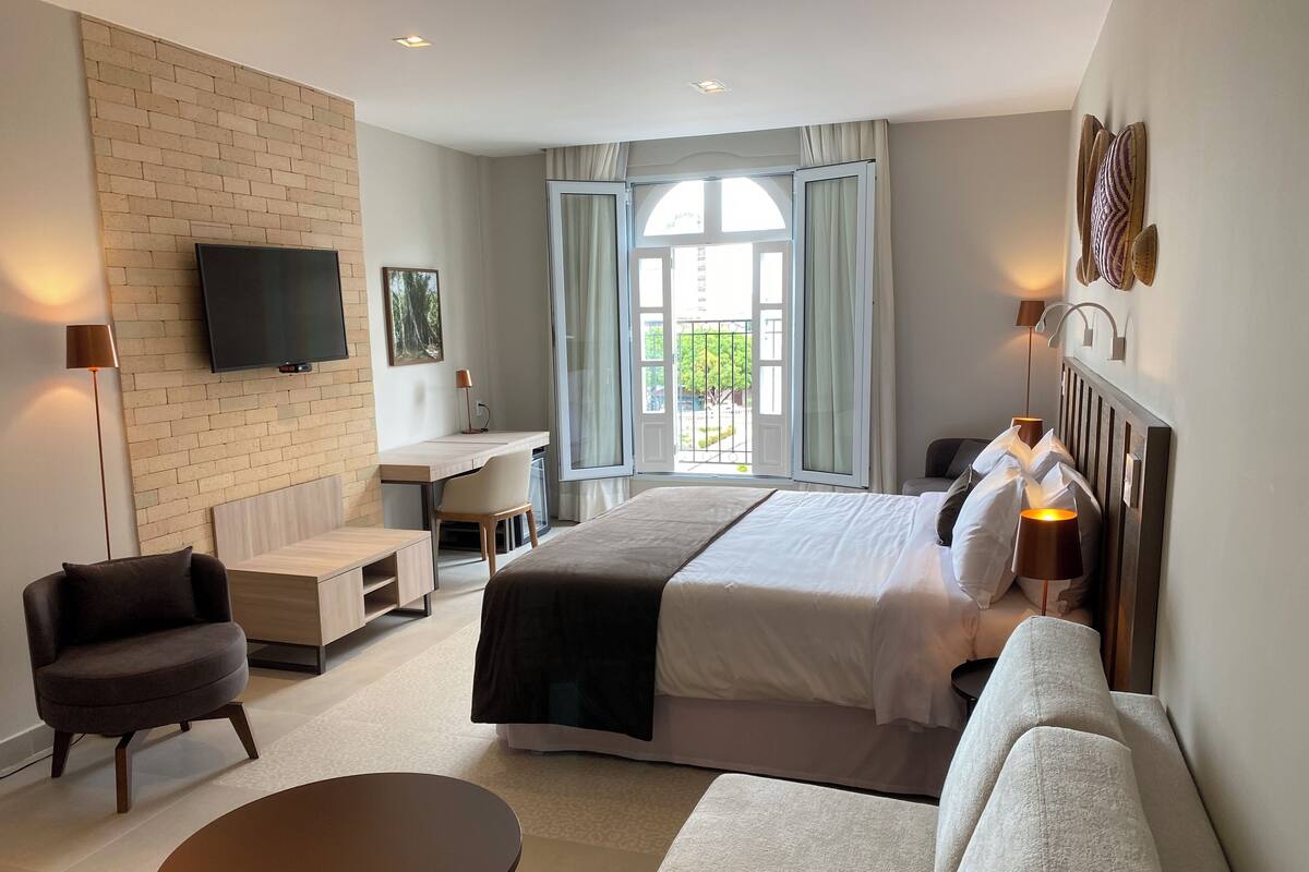 A modern hotel room with a large bed, a desk, a chair, a sofa, and a TV.