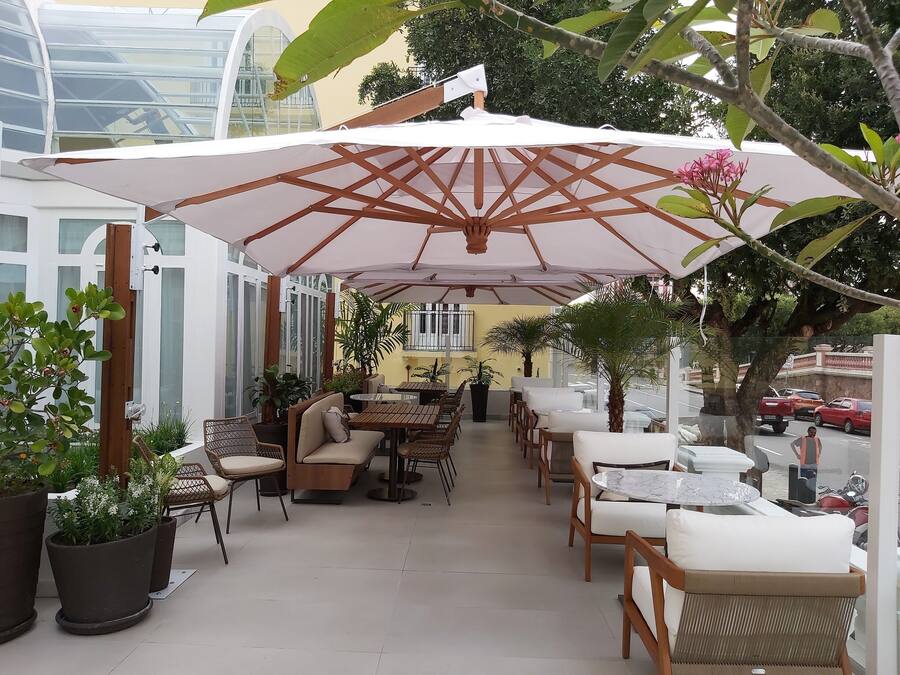 Un patio al aire libre con mesas y sillas de madera, cubierto por un gran paraguas, rodeado de plantas en macetas y un edificio de vidrio al fondo.
