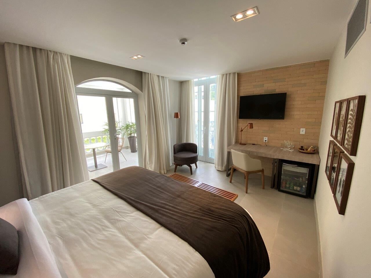 Ein Hotelzimmer mit einem großen Bett, einem Schreibtisch, einem Sessel, einem Fernseher und einem Balkon mit Vorhängen.