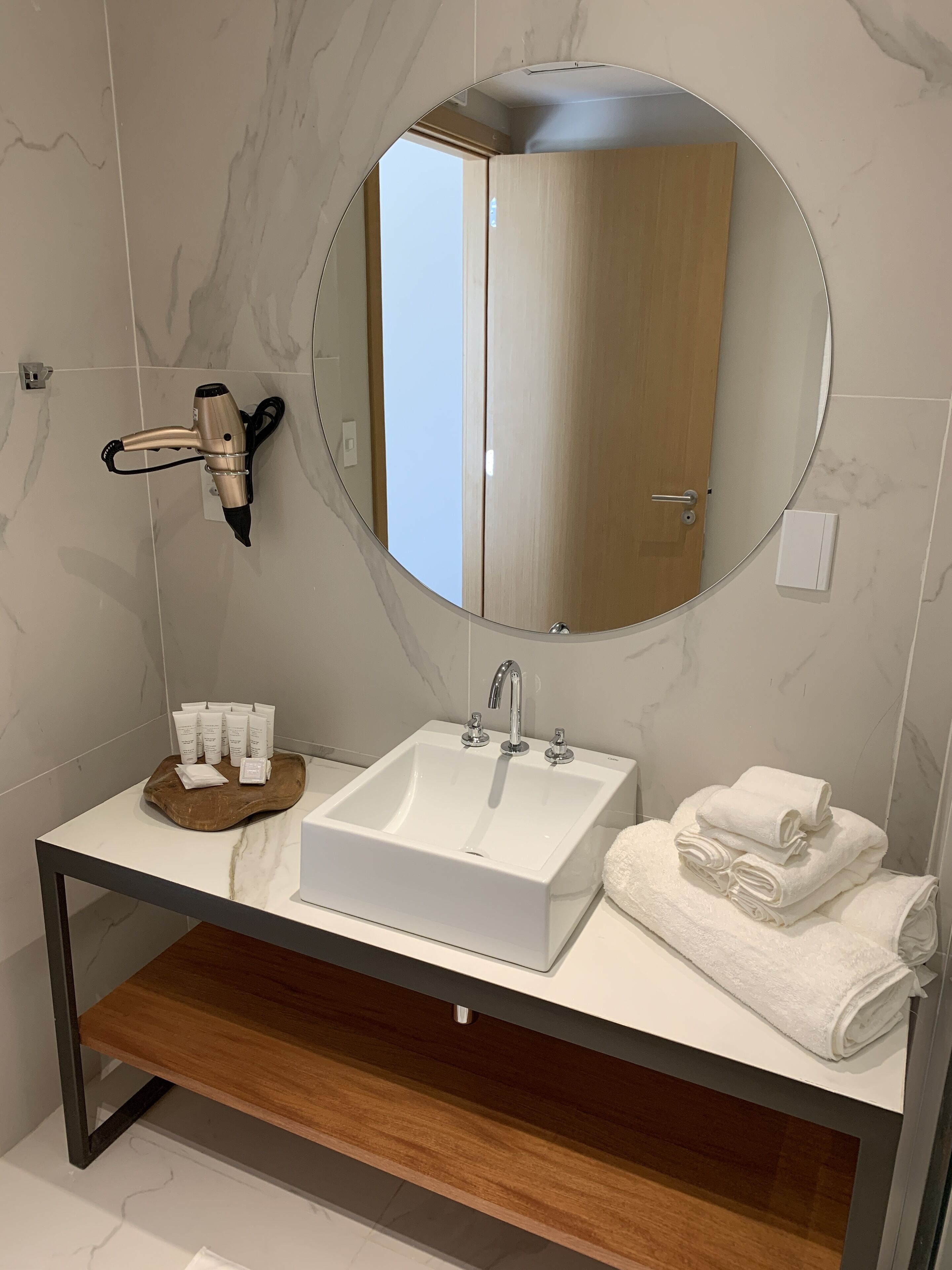 A modern bathroom with a white sink, a round mirror, and a hairdryer mounted on the wall.