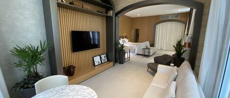 Habitación de hotel moderna con una mesa de comedor de mármol, un sofá, un televisor montado en una pared de madera y una cama al fondo.
