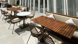 Un patio al aire libre con mesas de madera y sillas de mimbre, un área con techo curvo y paredes de vidrio, y un edificio amarillo con detalles blancos al fondo.