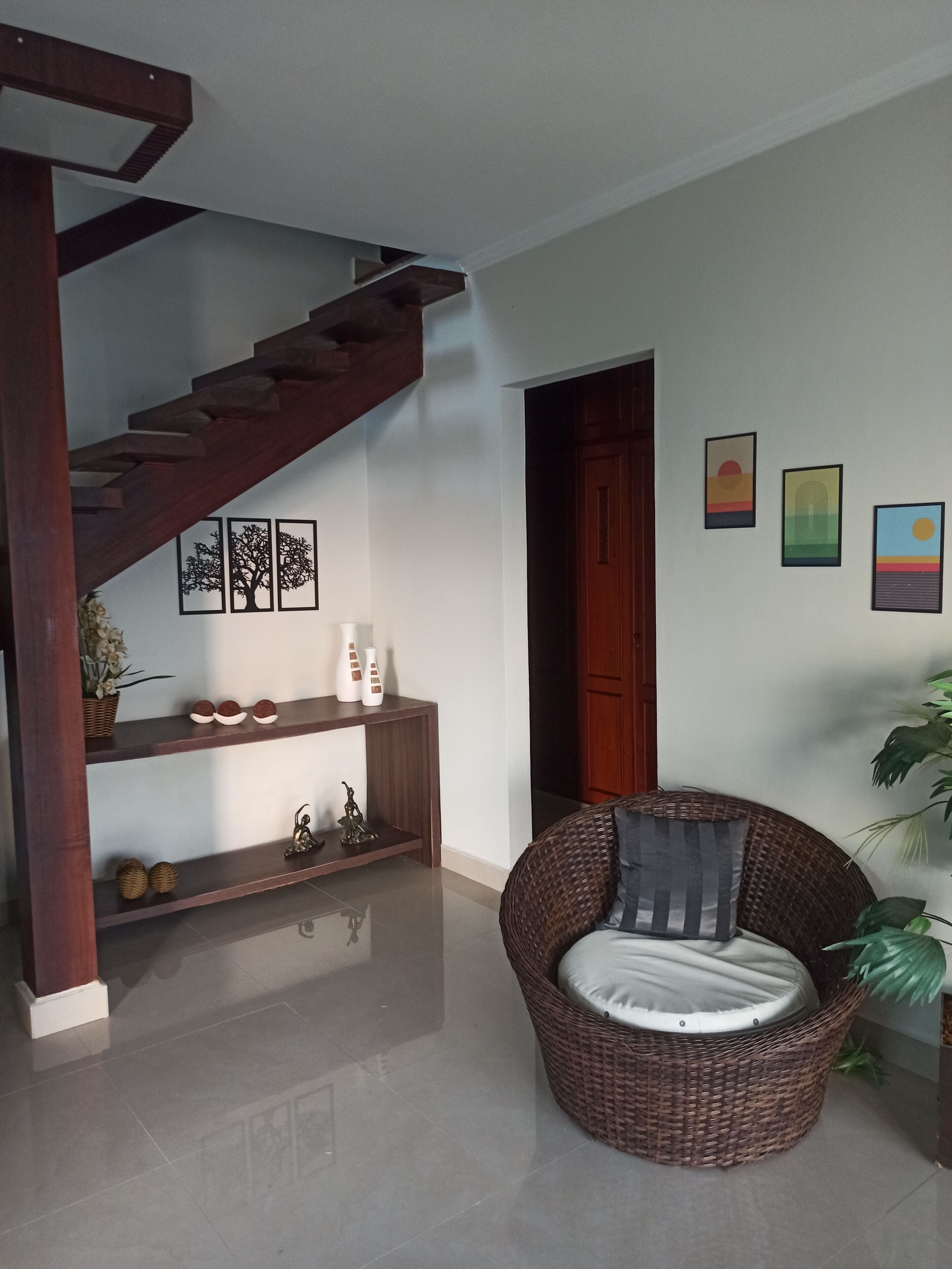 Uma sala de estar com escada de madeira, uma poltrona de vime com almofada, um console com objetos decorativos e três obras de arte emolduradas na parede.