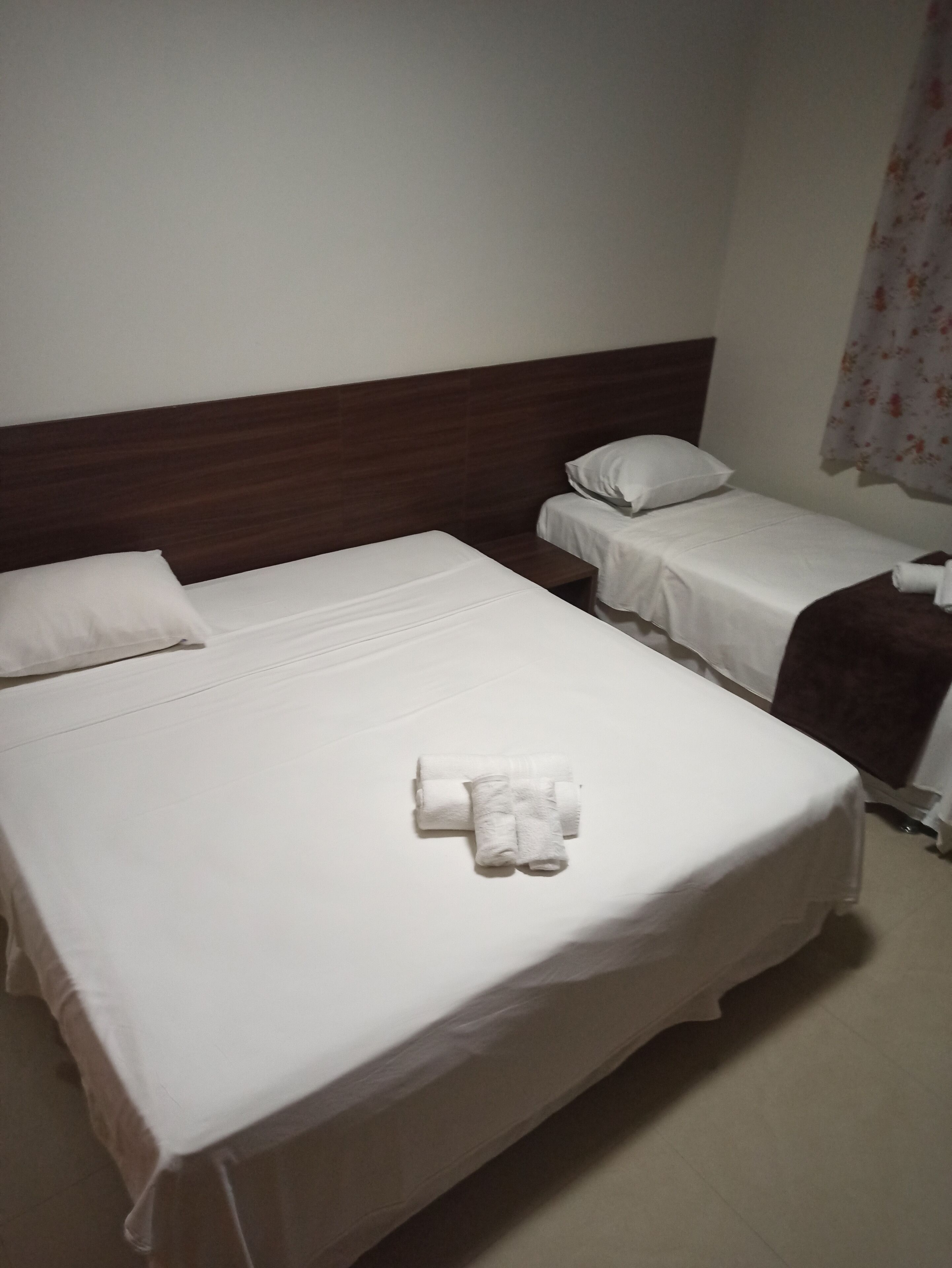Habitación de hotel con dos camas individuales, cabecera de madera, ropa de cama blanca y una toalla enrollada sobre la cama.