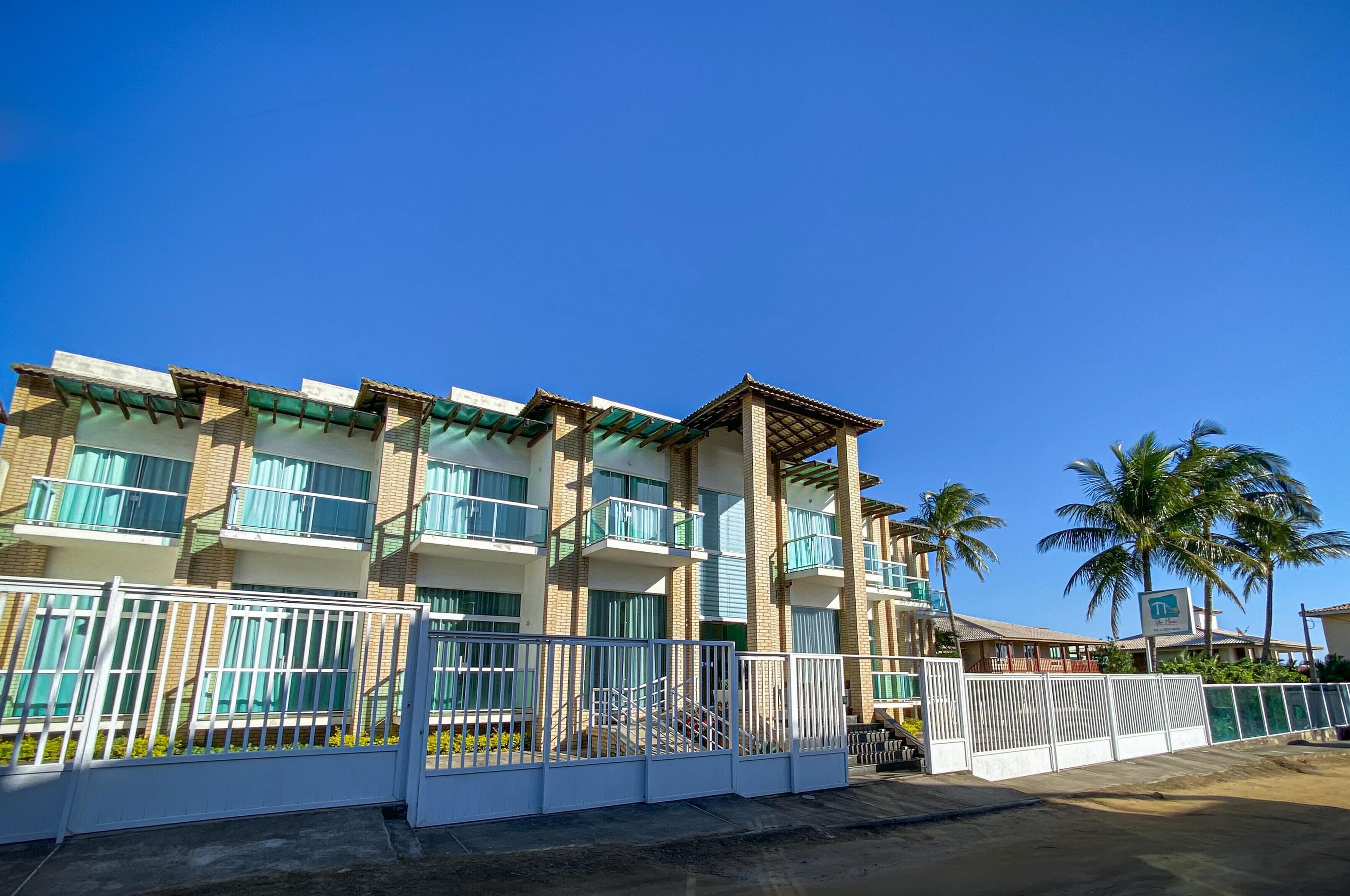A multi-story hotel with balconies, a gated entrance, and palm trees in the background.