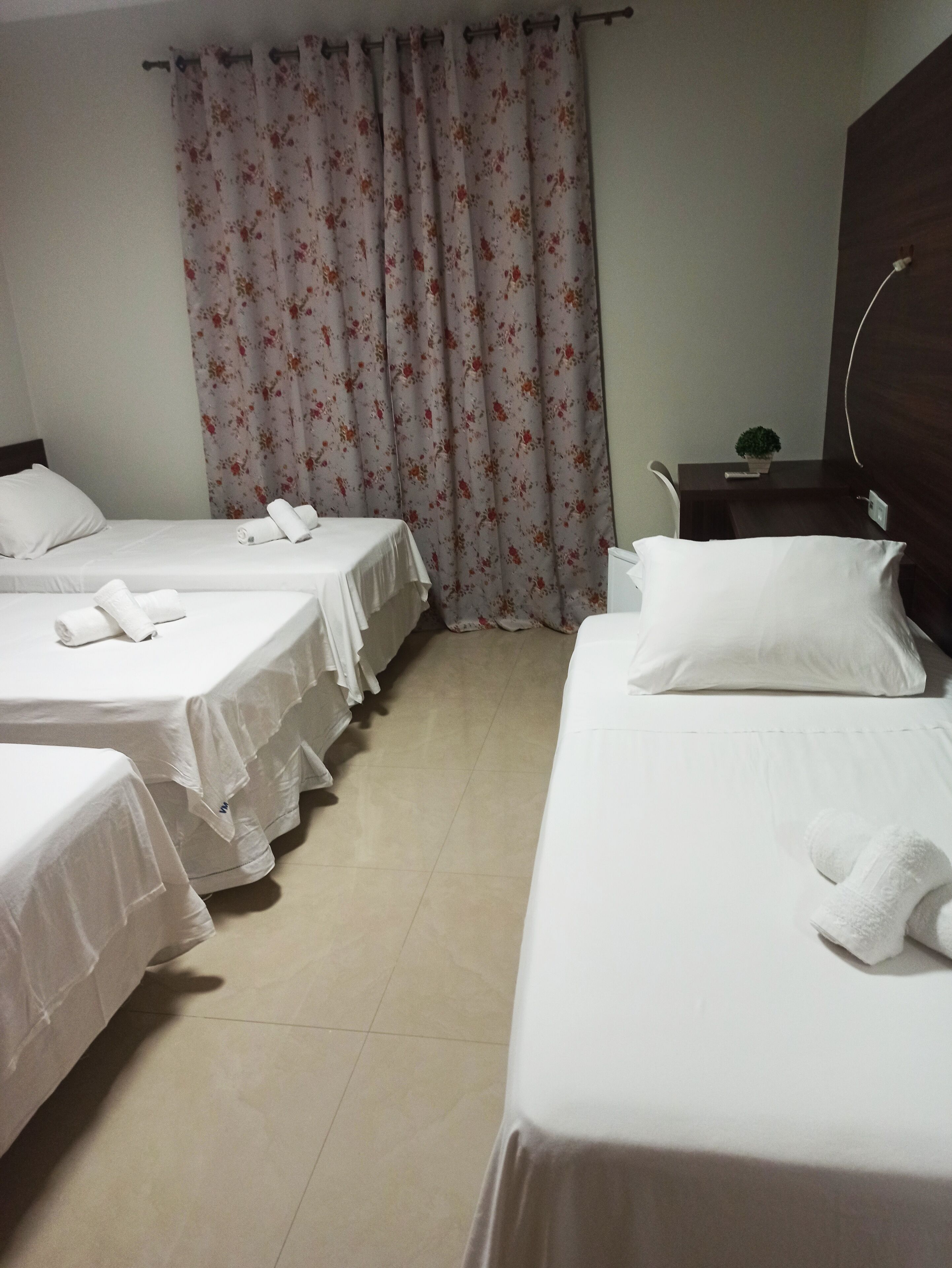 Habitación de hotel con tres camas individuales, cada una con una colcha blanca y toallas enrolladas. Hay una mesita de noche pequeña con una planta y un enchufe.