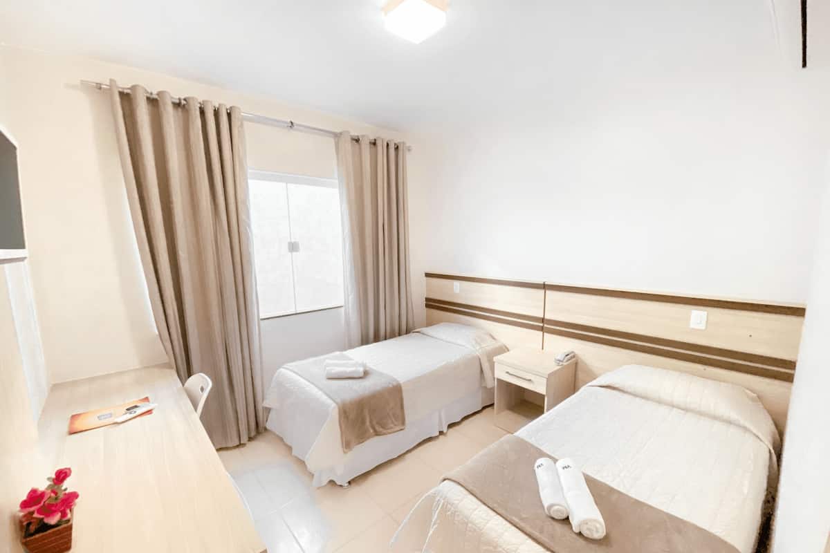Ein Hotelzimmer mit Bett, Nachttisch, Schreibtisch, Stuhl, einem Fenster mit Vorhängen und einer kleinen Pflanze.