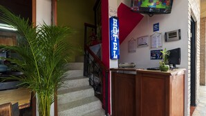 Un vestíbulo de hotel con una escalera, una mesa de recepción, una planta en maceta y una pantalla de televisión.
