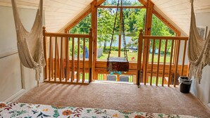 A room with a sloped ceiling, wooden railings, a hammock, a hanging swing, and a view of a lake and greenery outside.