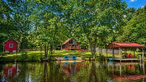 A lakeside cabin with a dock, a red barn, and a wooden deck with chairs.
