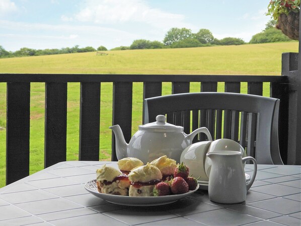 A table with a plate of scones and strawberries, a teapot, and a jug on a balcony overlooking a green field.