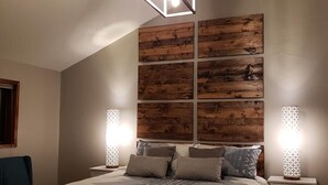 Ein modernes Schlafzimmer mit schräg verlaufender Decke, einer geometrischen Pendelleuchte, einer Holz-Akzentwand, einer gemusterten Tagesdecke und Nachttischlampen.
