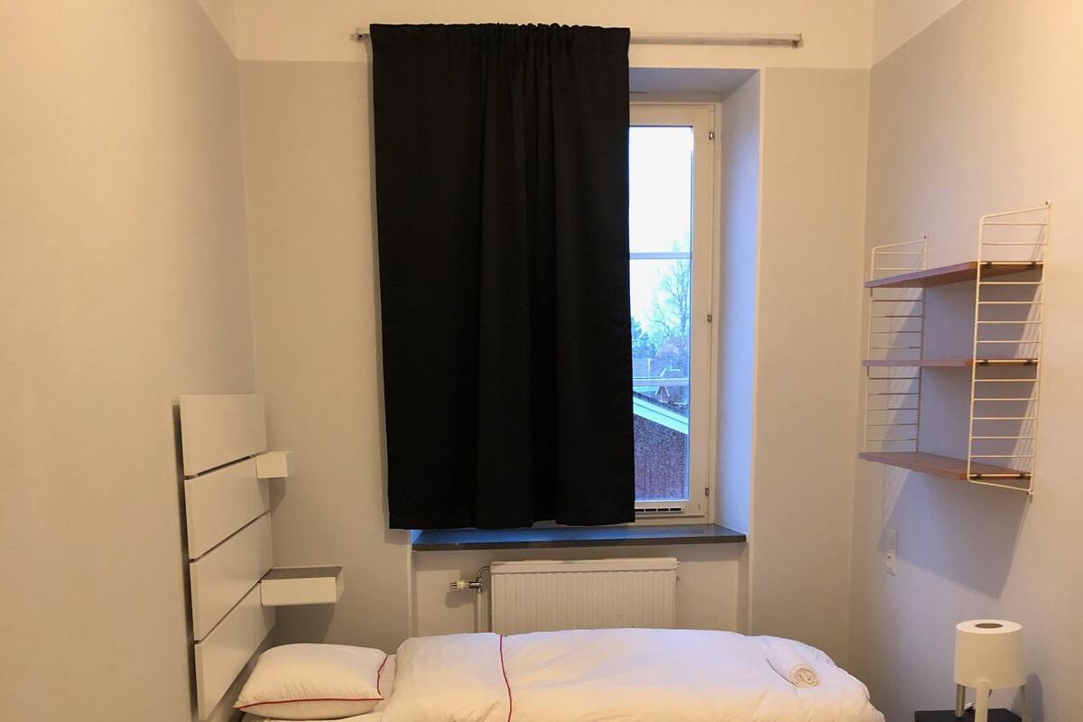 A small, neatly arranged bedroom with a single bed, a window with black curtains, a radiator, and a small table with a lamp.
