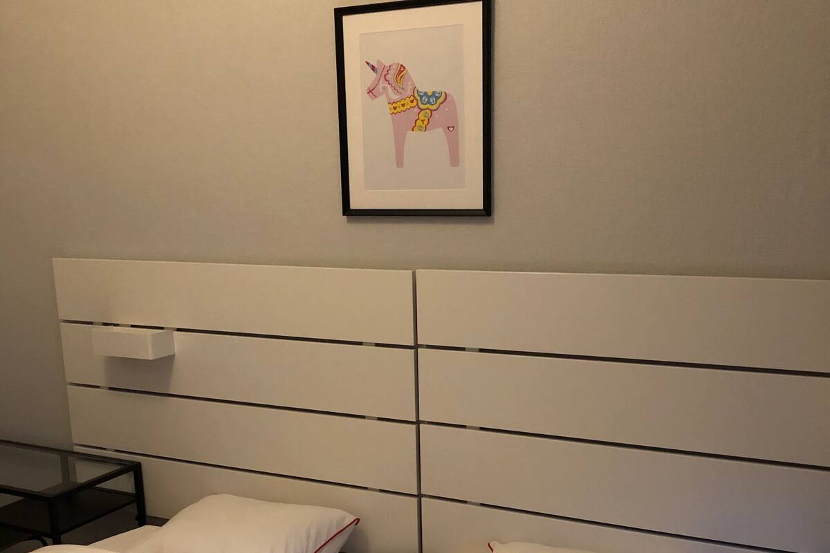A hotel room with a white headboard, two beds with white bedding and red piping, a small bedside table, and a framed picture of a unicorn on the wall.