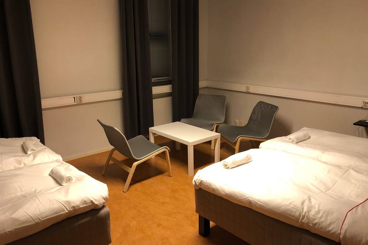 A hotel room with two beds, a small table, two chairs, a window with curtains, and a wall-mounted light fixture.