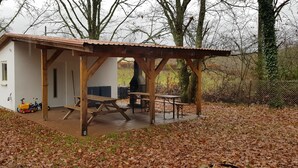 A wooden pavilion with a tiled floor and picnic tables, surrounded by trees and a fence.