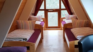 A triangular attic bedroom with two beds, a chair, and a window providing a view of trees.