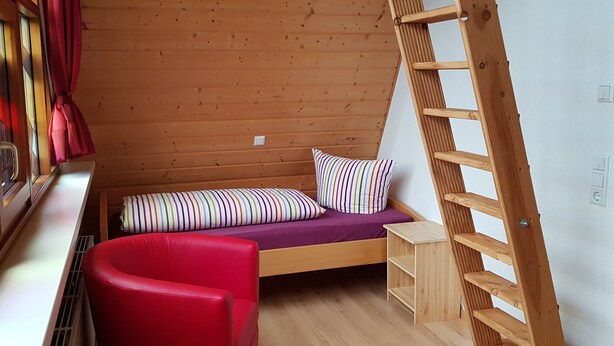 A small cabin with a wooden interior, a bunk bed with striped pillows, a red chair, and a wooden ladder.