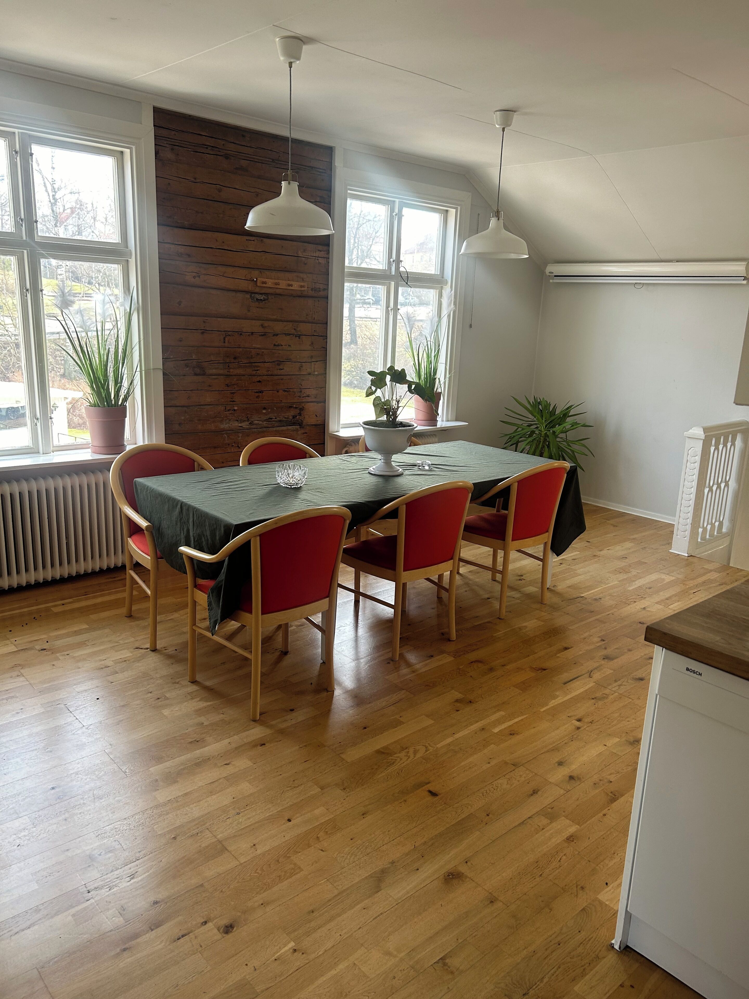 Uma área de refeições com mesa de madeira, cadeiras vermelhas, plantas em vasos e luminárias pendentes.