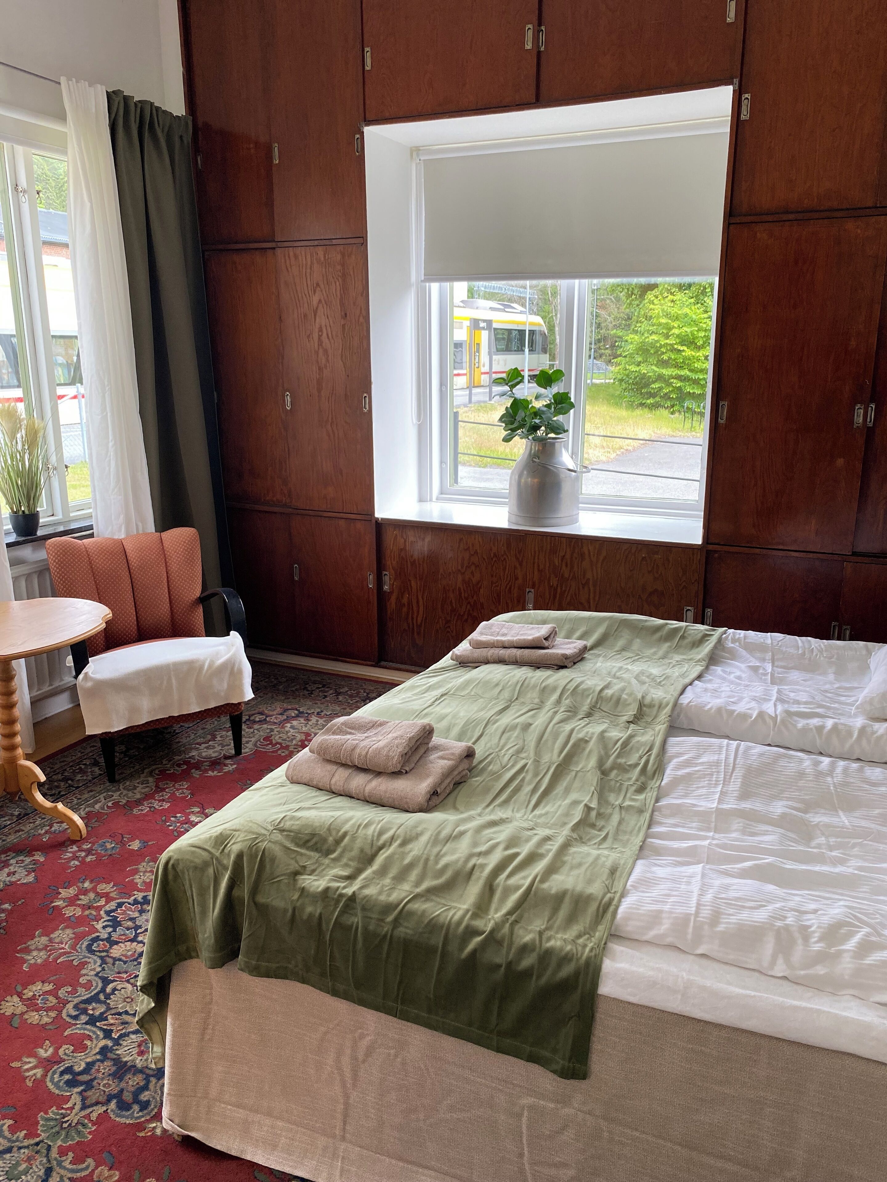 Un dormitorio con una cama, una silla, una mesita, una ventana con vista a un autobús y una planta en maceta en el alféizar.