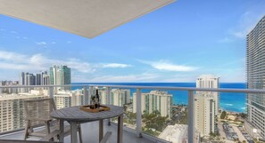 A balcony with a wooden table and chairs, overlooking a cityscape and the ocean.