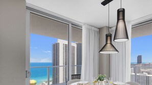 Comedor con una mesa redonda preparada para cuatro personas, ventanas amplias con vista a la ciudad y al océano, y dos lámparas colgantes.