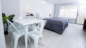 A modern hotel room with a white bed, a desk with a chair, a small table, and a potted plant. - Orlando Vacation Rooms (Kissimmee)