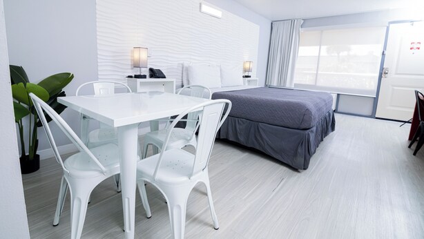A modern hotel room with a white bed, a desk with a chair, a small table, and a potted plant. - Orlando Vacation Rooms (Kissimmee)