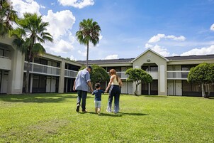 A family walking on a grassy lawn in front of a building with multiple balconies and palm trees.