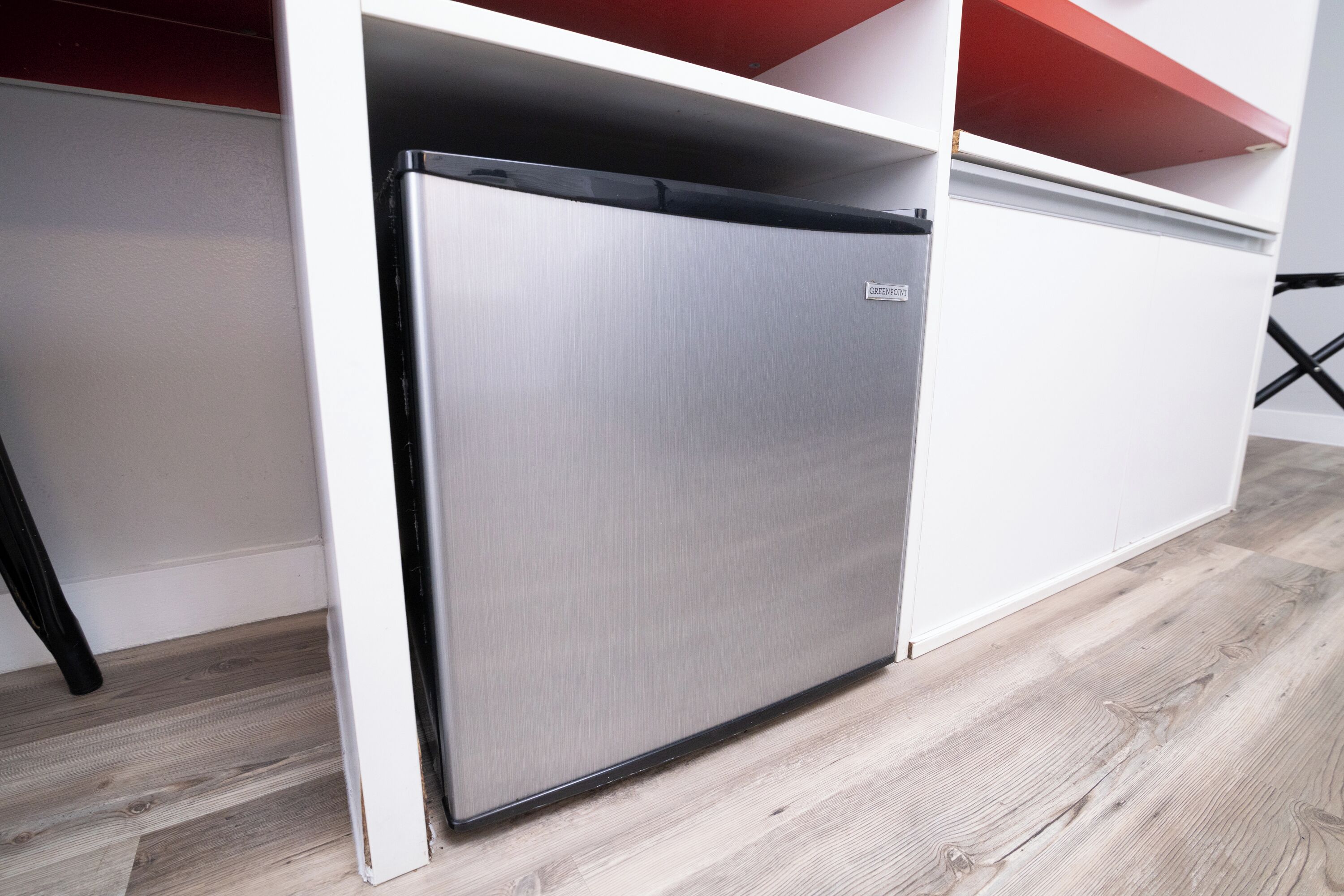 A compact, stainless steel refrigerator in a kitchen with a light wood floor and white cabinetry.