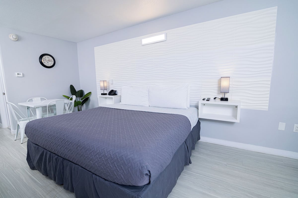 A modern bedroom with a large bed, a desk, and a chair. There is a clock on the wall and a plant on the desk.