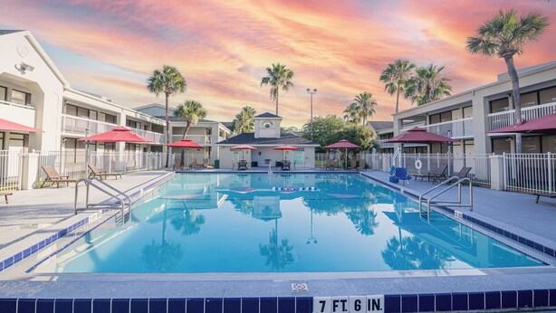 A swimming pool with a 7 ft. 6 in. depth, surrounded by palm trees and a building with red awnings. - Orlando Vacation Rooms (Kissimmee)