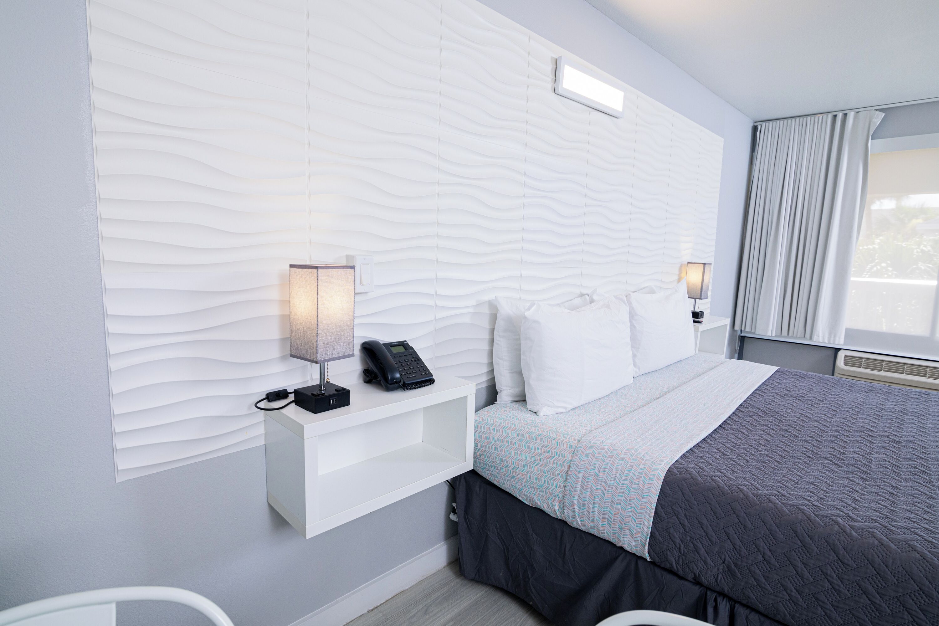 A modern hotel room with a white textured wall, a bed with a grey and blue patterned bedspread, a white bedside table with a telephone, and a nightstand with a lamp.