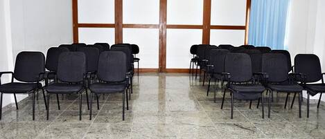 A conference room with rows of black chairs, a large window with wooden frames, and a tiled floor.