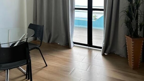 Una habitación moderna con una mesa de vidrio, dos sillas, una planta en maceta y vistas al exterior a través de amplios ventanales con cortinas grises.